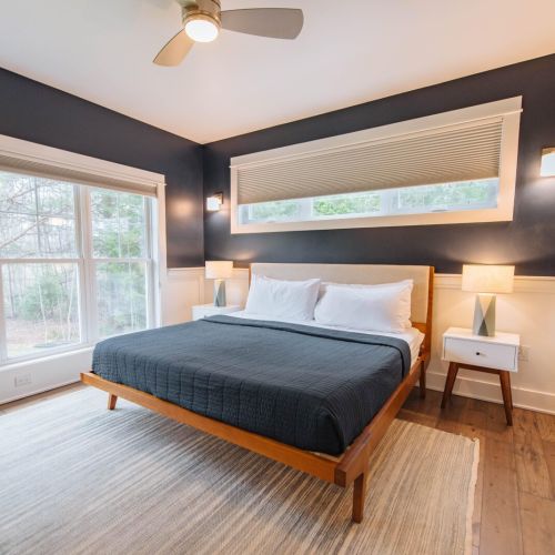 This spacious master bedroom features a luxurious king-sized bed, set against a sophisticated dark wall that enhances its grandeur. Large windows flood the room with natural light, offering serene views of the outdoors.