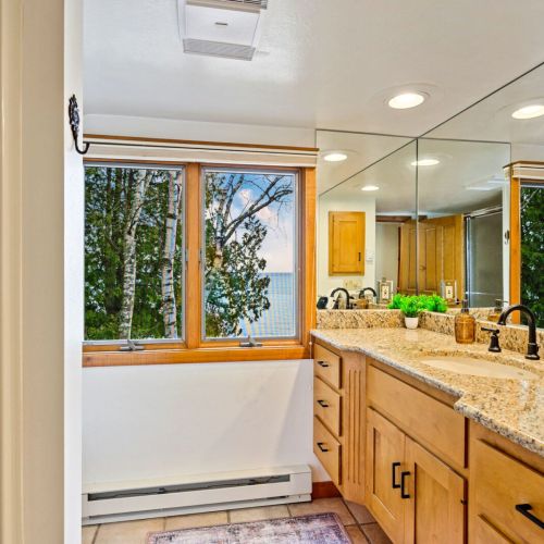 The large vanity area in the upper-level bathroom at Sandvik House provides ample space for your personal care routines, complemented by a generous mirror that reflects the natural light streaming through the window, offering views of the lake.