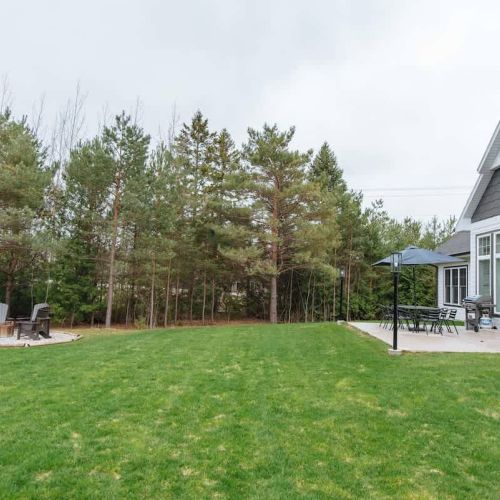 This expansive backyard features a well-manicured lawn that seamlessly transitions into a cozy outdoor living area. The patio, adjacent to the house, is equipped with a dining set under a large umbrella, perfect for alfresco meals. Nearby, a charming