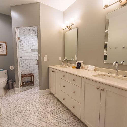 Step into this exquisite master bathroom, featuring high-end Kohler fixtures that blend functionality with elegance. The zero-entry shower provides a seamless transition, making it both accessible and stylish with its white subway tile. Dual vanities with ample storage ensure personal space for each user, while the hexagonal floor tiles add a touch of classic sophistication. Modern sconces and large mirrors complete the look, creating a spa-like atmosphere perfect for unwinding.